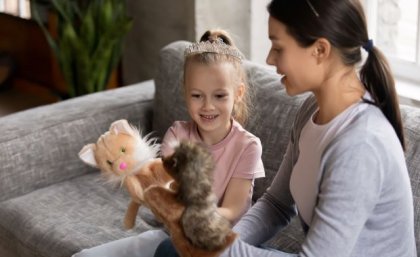 a child and a woman sit on a grey couch holding two animal puppets